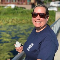 A white woman wears brown sunglasses and a dark blue shirt with the insignia of the City of Bloomington on the right side. The background is a pond covered in green vegetation 