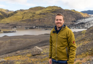 A white man with a brown mustache and wearing a greenish yellow sweater stands in front of a mountain backdrop 